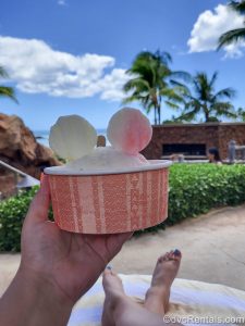 A hand holds up a red and pink cup holding a Mickey-shaped serving of shaved ice. The beach, blue sky, and palm trees can be seen in the background.