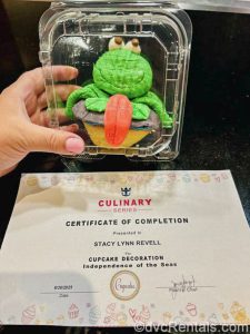 A cupcake decorated with a green fondant frog in seen in a plastic container along with a certificate of completion for a cupcake decorating class.