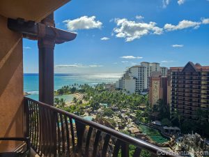 The ocean, beachfront, pool areas and exterior of Aulani, a Disney Resort & Spa are seen from a Villa balcony under a bright and sunny blue sky.