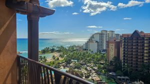 The ocean, beachfront, pool areas and exterior of Aulani, a Disney Resort & Spa are seen from a Villa balcony under a bright and sunny blue sky.