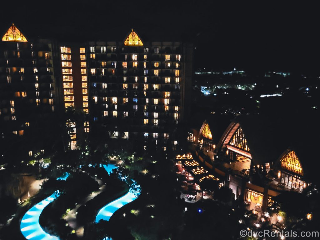 The lights of Aulani’s exterior and guest rooms at night are seen from a high floor Villa balcony. The pool area is also illuminated below.