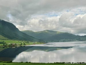 Scenic green fields and mountains are seen along the sides of the water under a cloudy sky near Molde, Norway.
