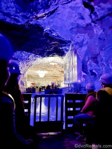 Passengers in hard hats sit on a wooden raft as it floats across the under-mountain waters in the Bergtatt Marble Caves. The interior rock formations of the mines are lit with purple and white lighting.