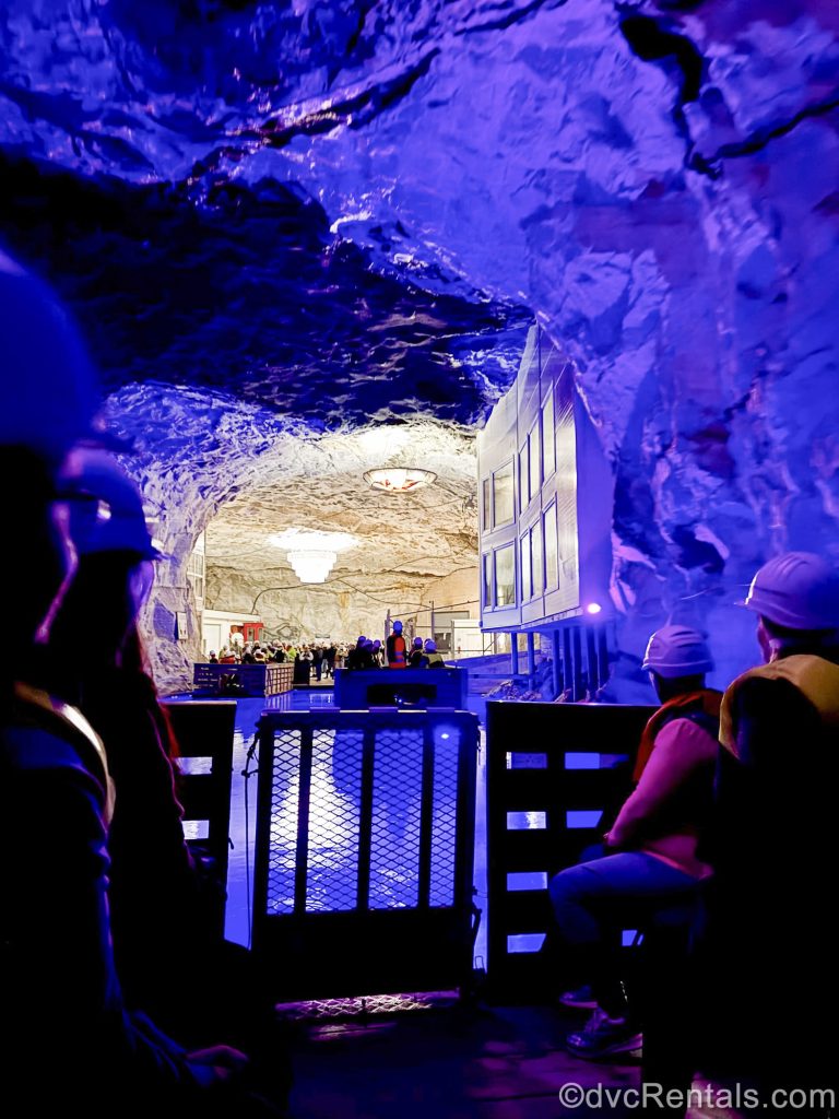 Passengers in hard hats sit on a wooden raft as it floats across the under-mountain waters in the Bergtatt Marble Caves. The interior rock formations of the mines are lit with purple and white lighting.