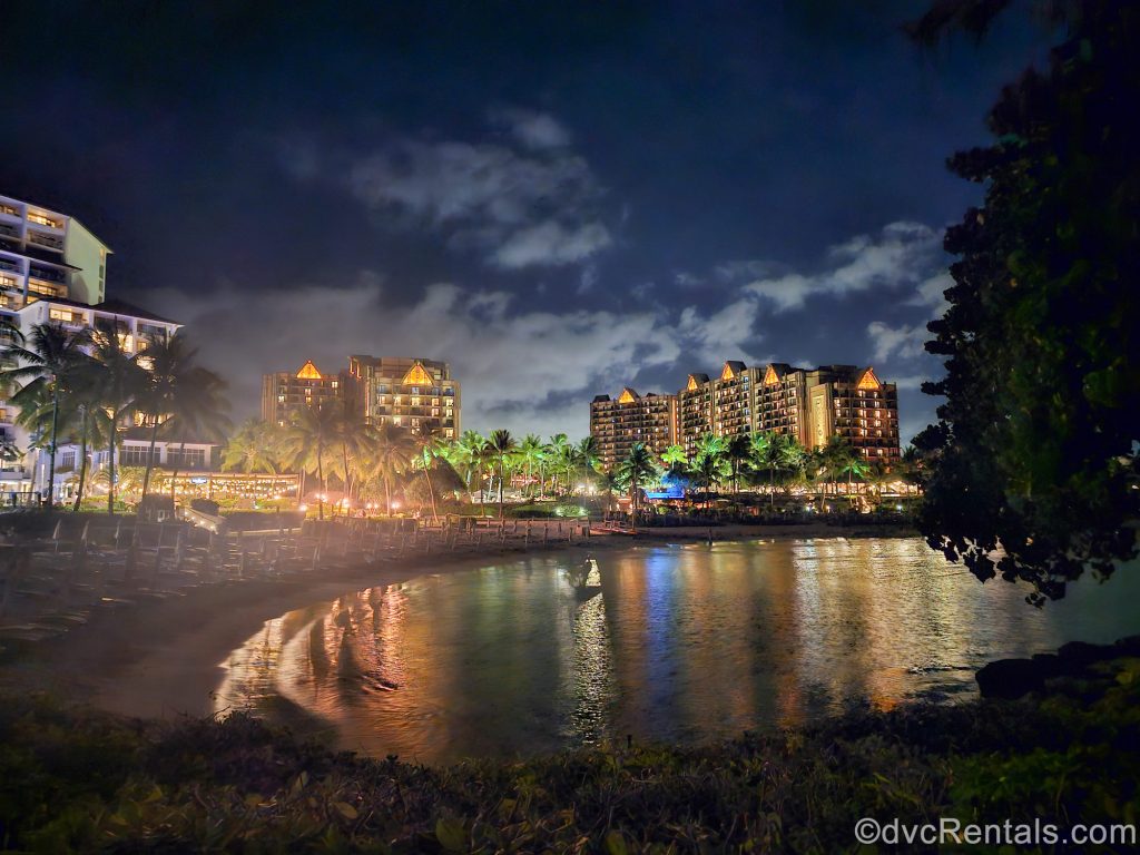 Aulani, a Disney Resort & Spa is seen from across the beach at night as the resort is illuminated with warm lighting.