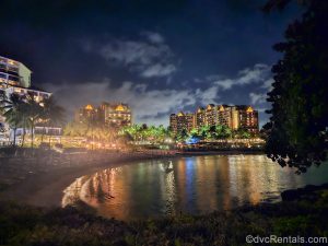 Aulani, a Disney Resort & Spa is seen from across the beach at night as the resort is illuminated with warm lighting.