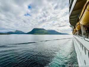 The white siding and portholes of a cruise ship are seen as it floats through a Norwegian Fjord with mountains in the distance.