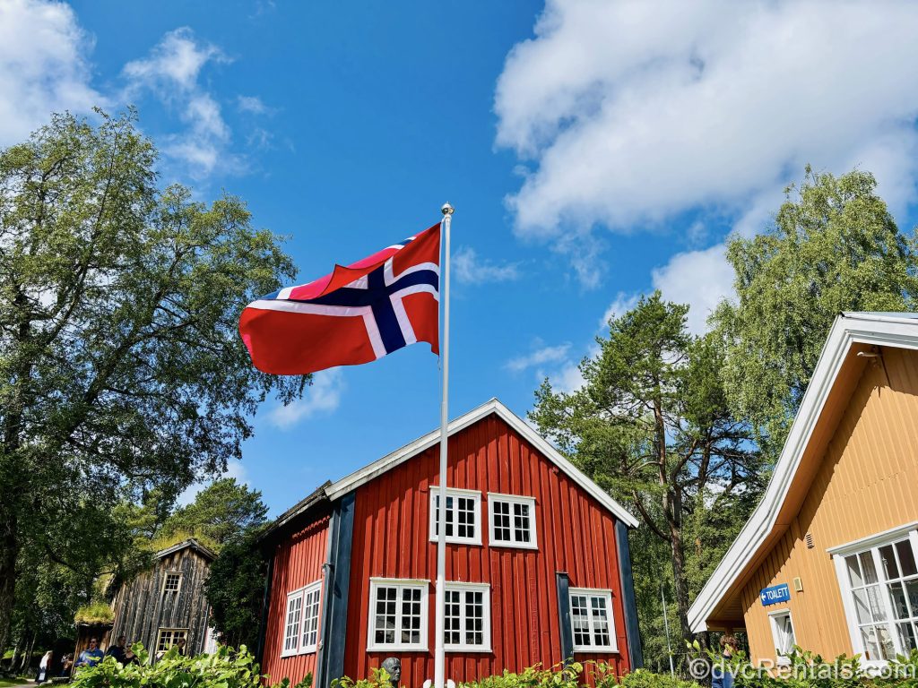 Red, yellow, and dark wood buildings of the Romsdal Museum in Molde, Norway are seen under a sunny sky and the Norwegian flag flaps in the breeze