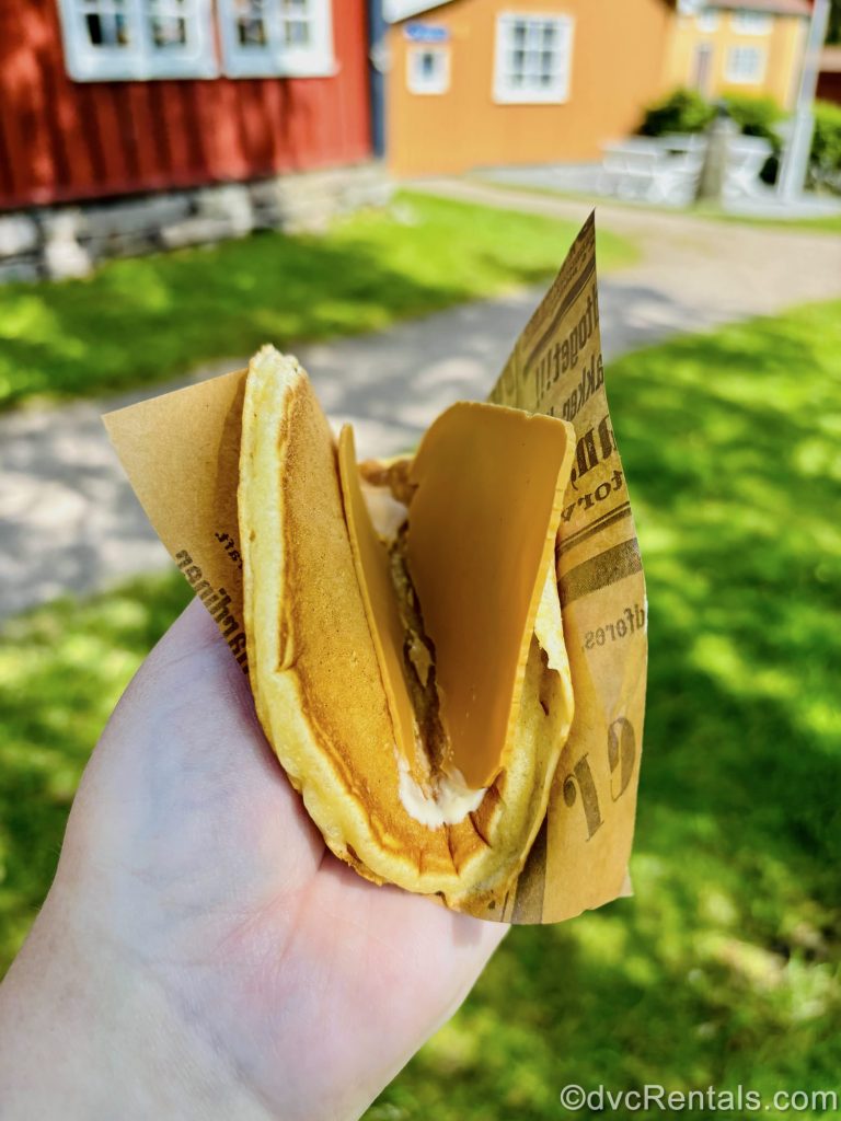 A hand holds a serving of svele, a Norwegian snack made of a pancake topped with brown cheese and butter that is folded like a taco.