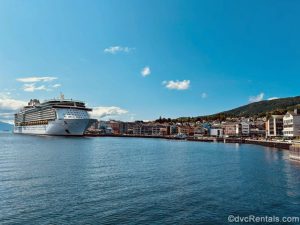 Royal Caribbean’s Independence of the Seas is seen docked along the waterfront of Molde, Norway under a sunny, blue sky. The town’s buildings can be seen, along with mountains in the distance.
