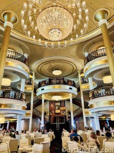 The main dining room of the Independence of the Seas. Guests are seated at tables with white chairs on multiple floors as a large, elegant crystal chandelier hangs overhead.
