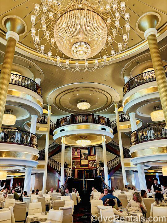 The main dining room of the Independence of the Seas. Guests are seated at tables with white chairs on multiple floors as a large, elegant crystal chandelier hangs overhead.