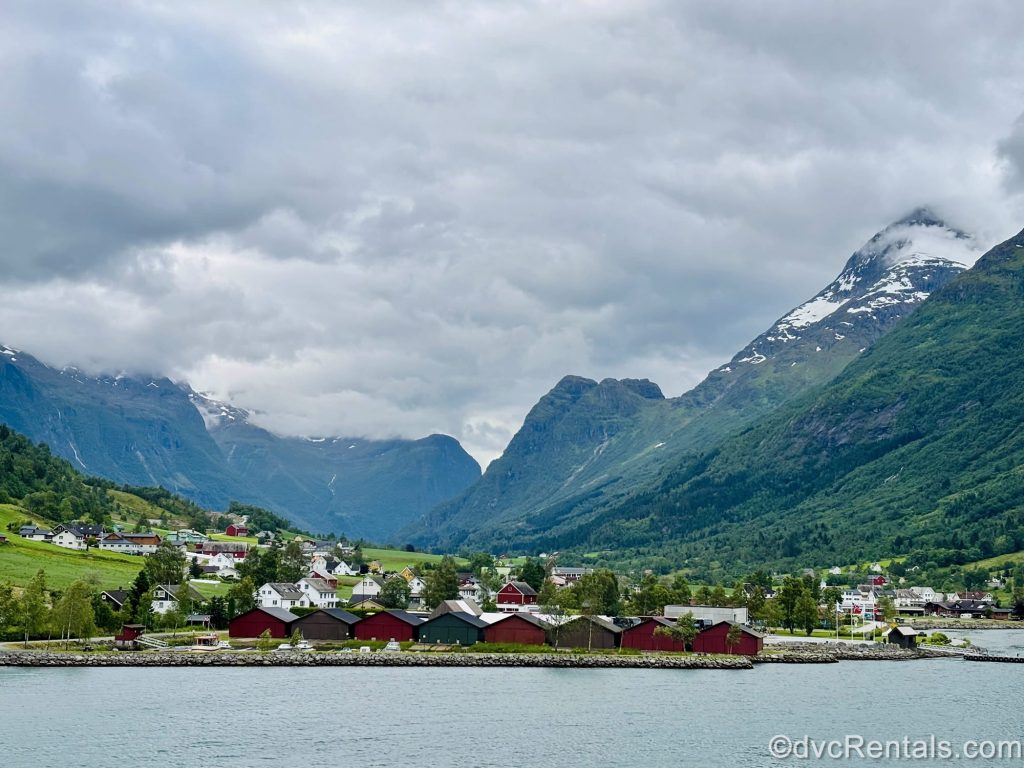 Mountains fill the background of a scenic view of Olden, Norway. There are white, red, and green buildings seen along with vivid green fields and the waters of the Nordfjord.
