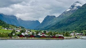 Mountains fill the background of a scenic view of Olden, Norway. There are white, red, and green buildings seen along with vivid green fields and the waters of the Nordfjord.