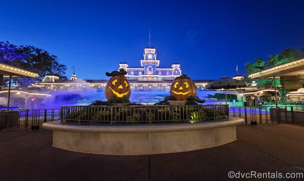 2 large jack-o’-lanterns are displayed in front of the entry to Magic Kingdom at night. There is blue fog and green lighting, as the park is decorated for Halloween.