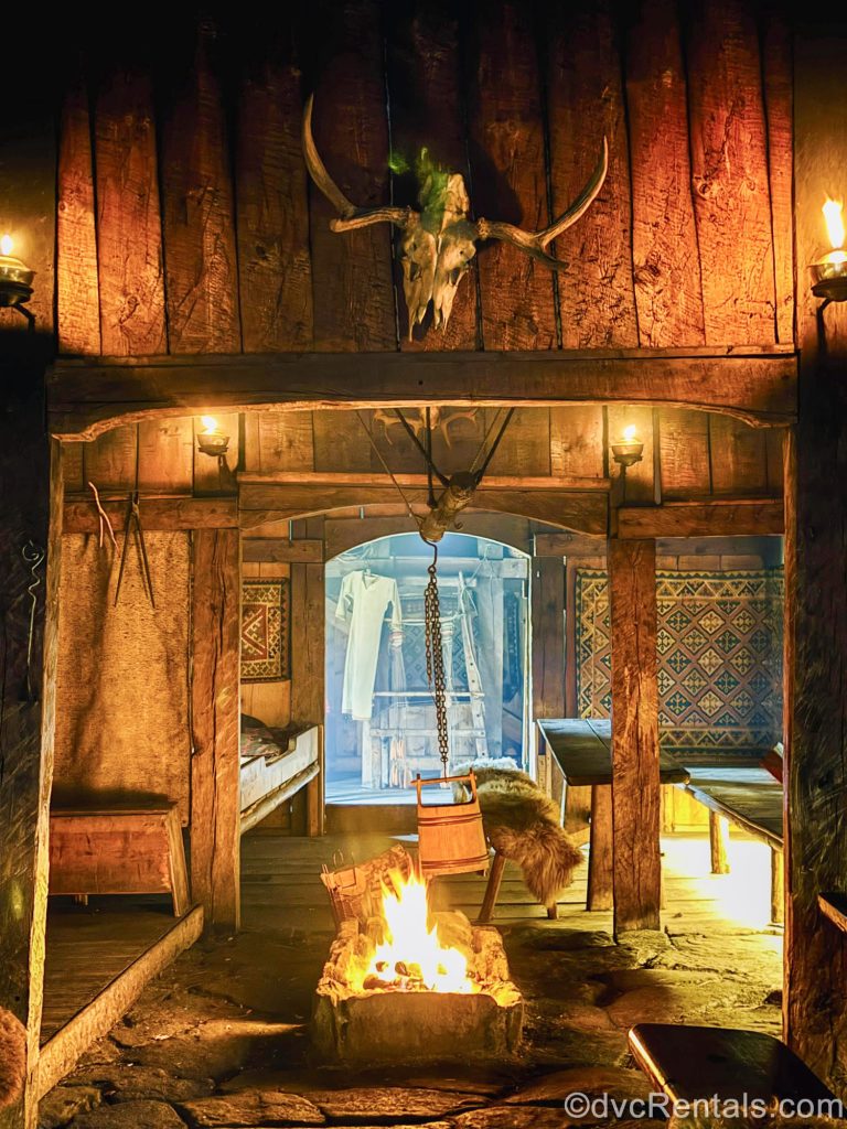 The wooden interior of a recreated Viking longhouse is lit by a large fire in the center of the room. Benches for seating and other artifacts from the time period are seen around the space.