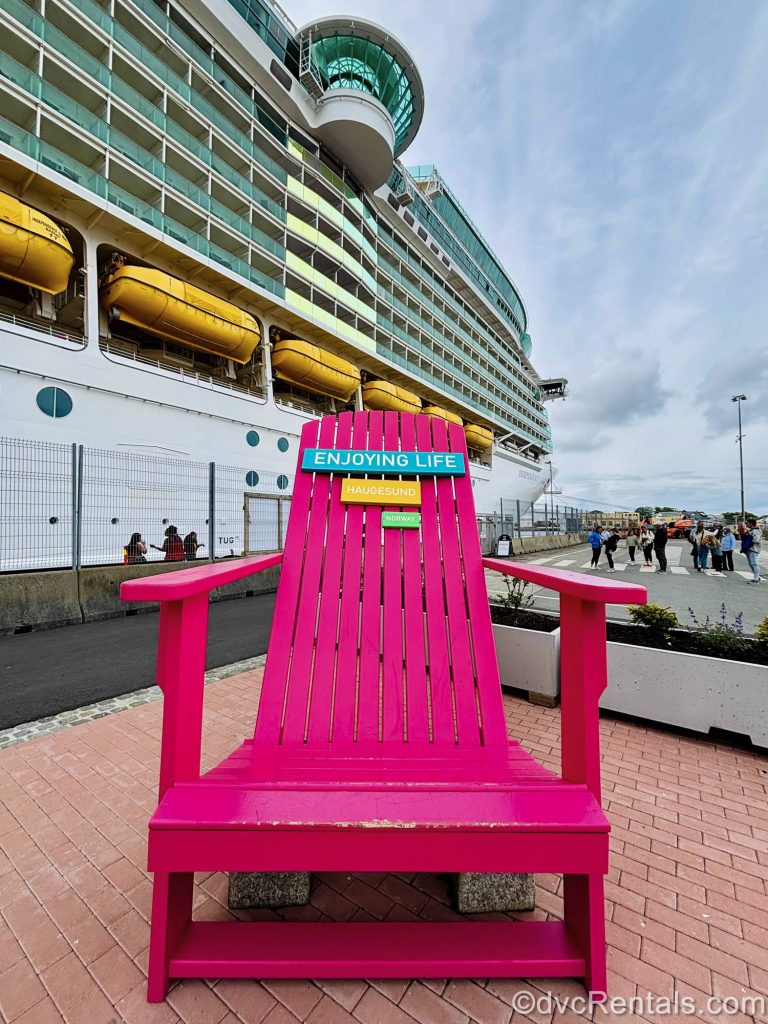 A pink, oversized deck chair with a sign reading “Enjoying life, Haugesund, Norway” is seen in front of Independence of the Seas as passengers board the ship in port in the background.