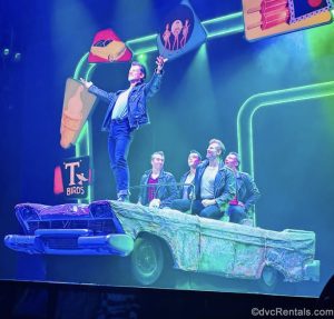 The cast members of Grease perform a number during the show. They’re dressed in denim and black leather jackets, as they sing and dance in a prop car on stage.