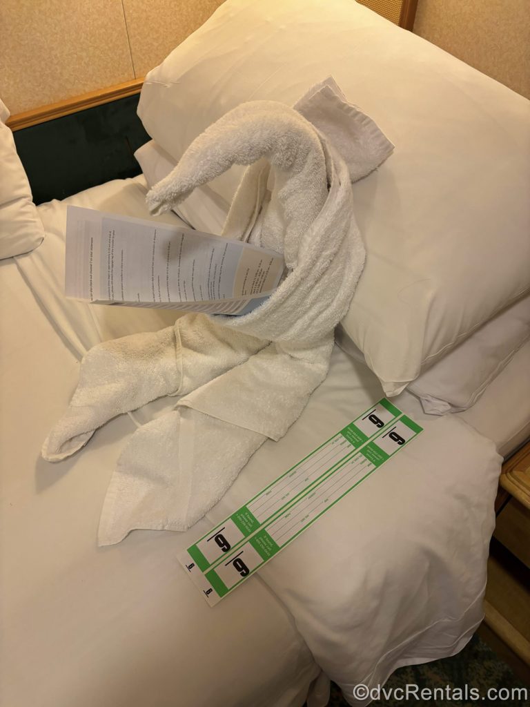 A white towel animal is arranged to look like it’s reading a form with cruise debarkation information on a stateroom bed, with green luggage tags on the side.