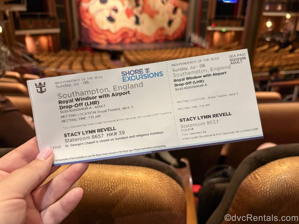 A hand holds up a cruise excursion ticket in the Royal Theater. The ticket is for a visit to Windsor Castle in England, followed by airport drop-off.