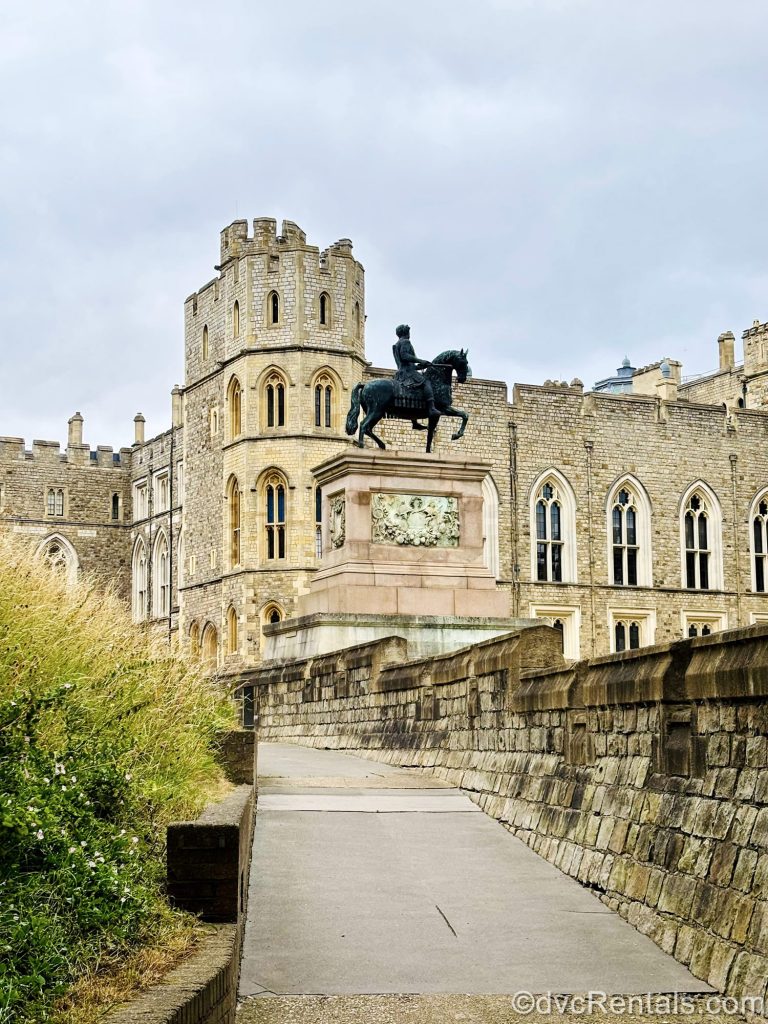 A statue of a rider on a black horse is seen in front of the light brown brick exterior of Windsor Castle in England.