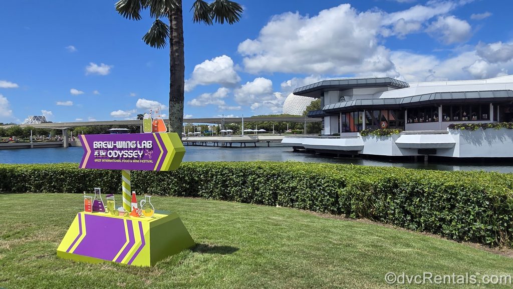 The purple and lime green sign for Brew-Wing at the Odyssey sits on the grass in front of the white building at EPCOT under a blue sky.