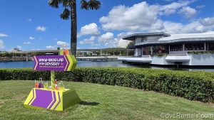 The purple and lime green sign for Brew-Wing at the Odyssey sits on the grass in front of the white building at EPCOT under a blue sky.