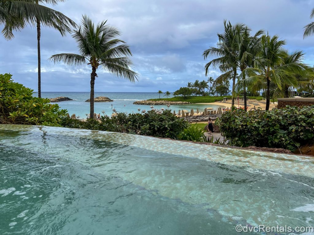 The beachfront at Aulani is seen from one of the resort’s pools under a blue sky. There are many loungers on the beach as palm trees tower above.