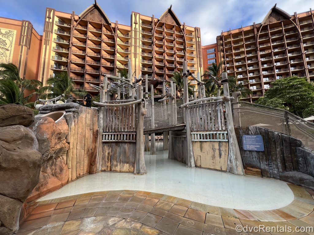 A water play area with a wooden footbridge at Aulani, a Disney Resort & Spa is empty in the early morning as golden sunlight shines on the resort exterior.