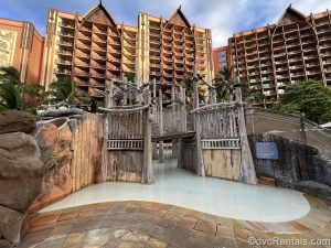 A water play area with a wooden footbridge at Aulani, a Disney Resort & Spa is empty in the early morning as golden sunlight shines on the resort exterior.