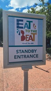 A white and blue sign reading “Eat to the Beat Standby Entrance” in colorful lettering is displayed at EPCOT during the Food & Wine Festival.