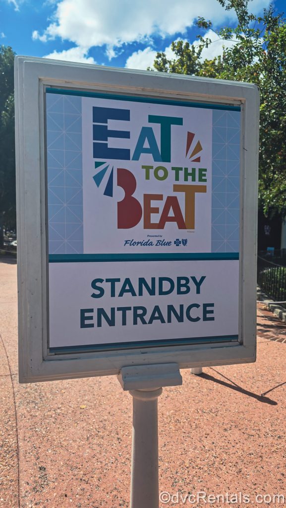 A white and blue sign reading “Eat to the Beat Standby Entrance” in colorful lettering is displayed at EPCOT during the Food & Wine Festival.