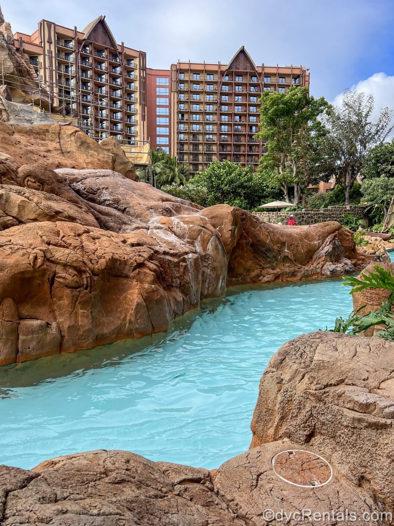 The lazy river at Aulani is seen under a blue sky with no guests yet in it. The turquoise water flows between brown rock walls with the resort exterior visible in the background.