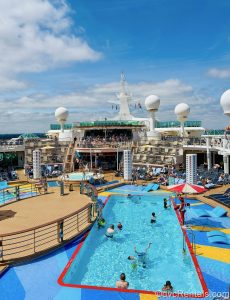 Cruise passengers enjoy the pools, hot tubs, and sun loungers on Independence of the Seas under a sunny, blue sky.