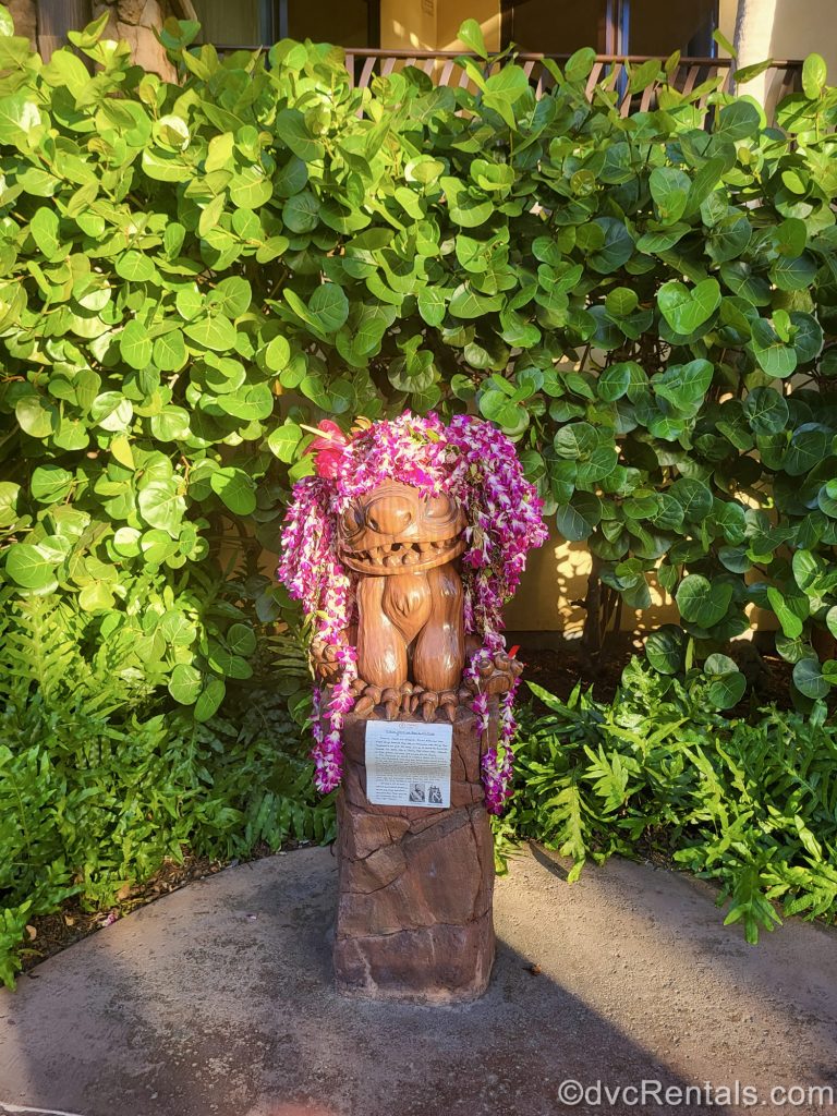 A wooden carving of Stitch is draped with pink flowers in front of some greenery.