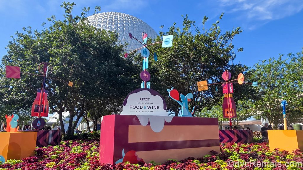 Colorful signage for the 2025 EPCOT International Food & Wine Festival featuring characters from Ratatouille are seen in front of the Spaceship Earth ball under a blue sky.