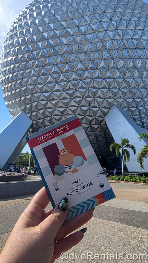 A hand holds up the 2025 EPCOT International Food & Wine Festival “passport” booklet in front of Spaceship Earth.