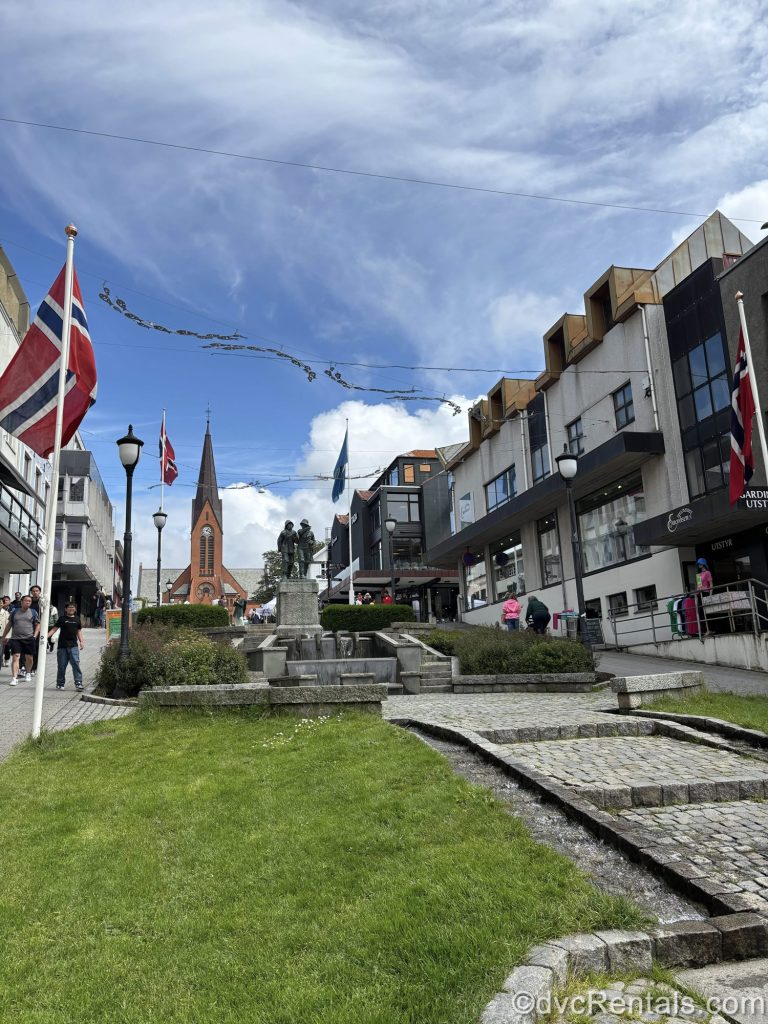A pedestrian street in Haugesund, Norway with a statue, shops, visitors, and the steeple of a church in the distance are seen under a blue sky