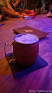The Cursed Treasure beverage in a copper mug sits upon the table with a menu in the background. The drink is topped with a white foam