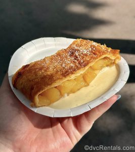 A hand holds up a serving of Apple Strudel with Vanilla Sauce on a white plate.
