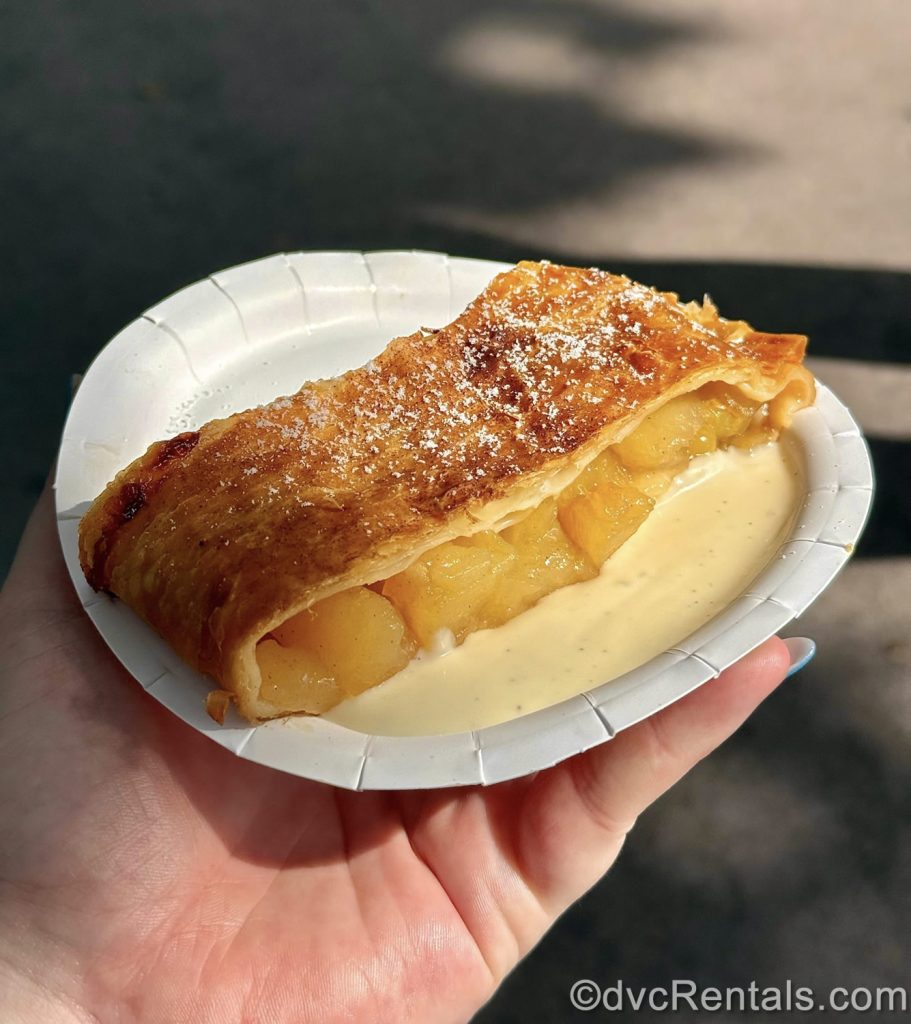 A hand holds up a serving of Apple Strudel with Vanilla Sauce on a white plate.