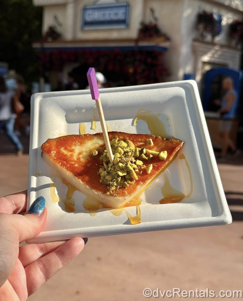 A hand holds up a serving of Griddled Cheese with Pistachios and Honey in front of the Greece booth at EPCOT.