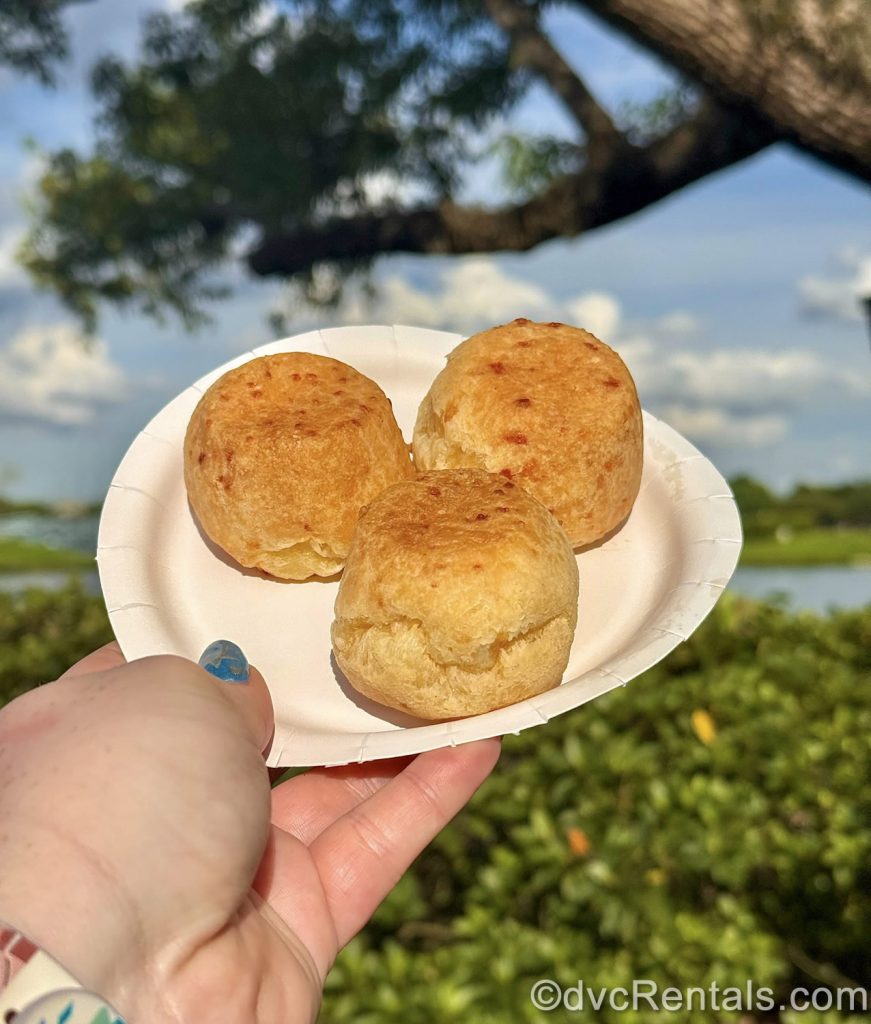 A hand holds up a serving of Pao de Queijo: Brazilian Cheese Bread in front of World Showcase Lagoon at EPCOT.