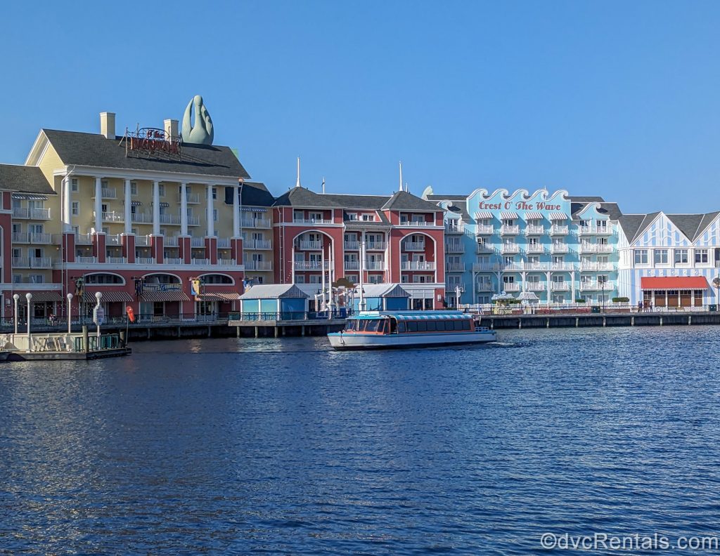 A white and blue Friendship Boat sails over Crescent Lake past Disney’s BoardWalk Villas under a sunny sky.