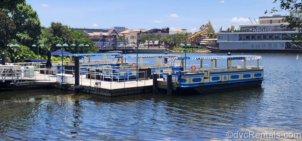 Three yellow and blue water taxis are docked outside Disney Springs, ready to take guests to nearby resorts Saratoga Springs and Old Key West. The restaurants and shops of Disney Springs are visible in the background under a bright, sunny sky.