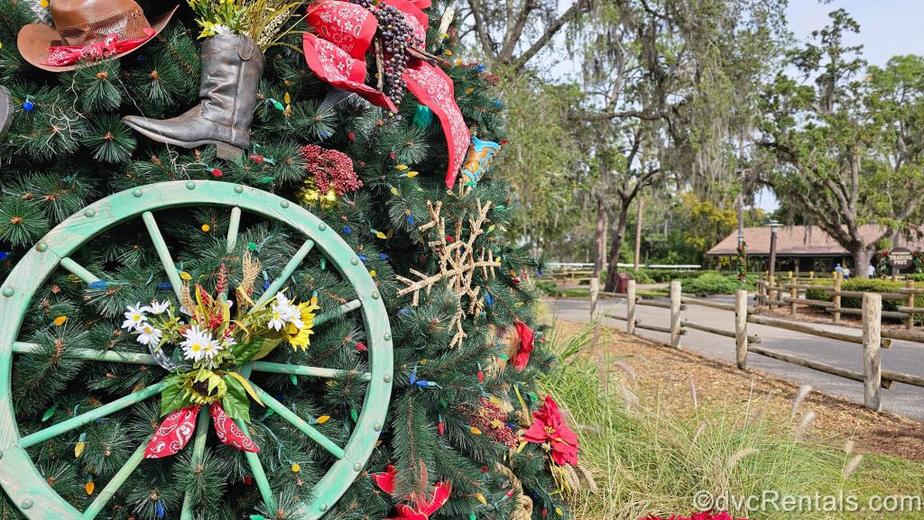 A large Christmas tree decorated with brown, red, and green frontier-themed ornaments like cowboy hats and wagon wheels is displayed outdoors at Disney’s Fort Wilderness Resort.