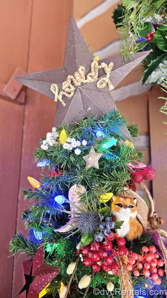 A small Christmas tree is decorated with frontier-themed ornaments like cowboy boots, stars, and animals. It is topped with a brown star that has the word “howdy” spelled out on it in brown rope.
