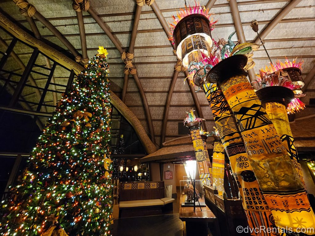A very large Christmas tree decorated with African-inspired ornaments and red and green lights. It is displayed in the lobby of Disney’s Animal Kingdom Villas Kidani Village, along with other African art pieces and artifacts.