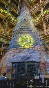 A wreath decorated with white lights and outdoor-theme baubles is displayed on the large stone fireplace in the lobby of Disney’s Wilderness Lodge. There are matching garlands draped above, as guests sit in wooden rocking chairs in front of the fire.