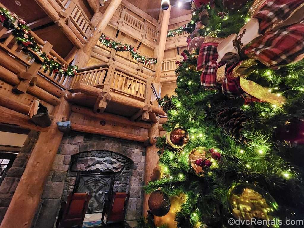 A green Christmas tree adorned with gold and red wilderness-themed ornaments is see in the lobby of the Boulder Ridge Villas at Disney’s Wilderness Lodge. Matching garlands are hung on the upper, wooden railings, and a fireplace with 2 chairs is seen in the background.
